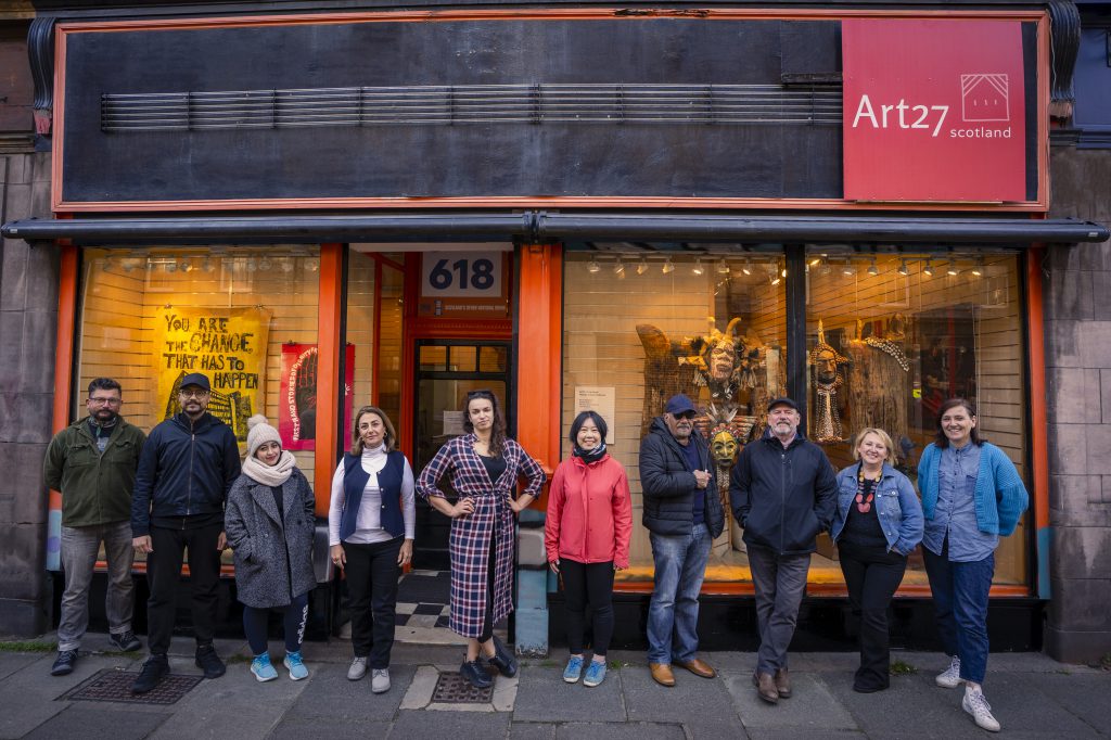 Ten people stand in a line, outside a shopfront which bears the logo of Art27.