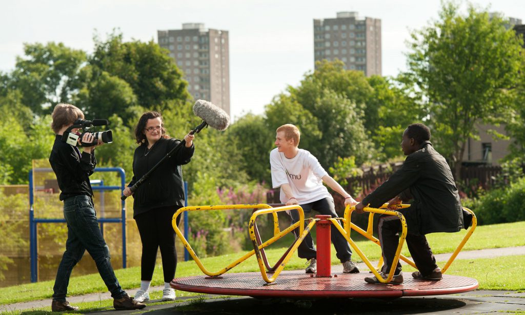 Four young people in a sunny park. One holds a video camera, another holds a boom microphone and the two others sit on a playpark carousel, being filmed.