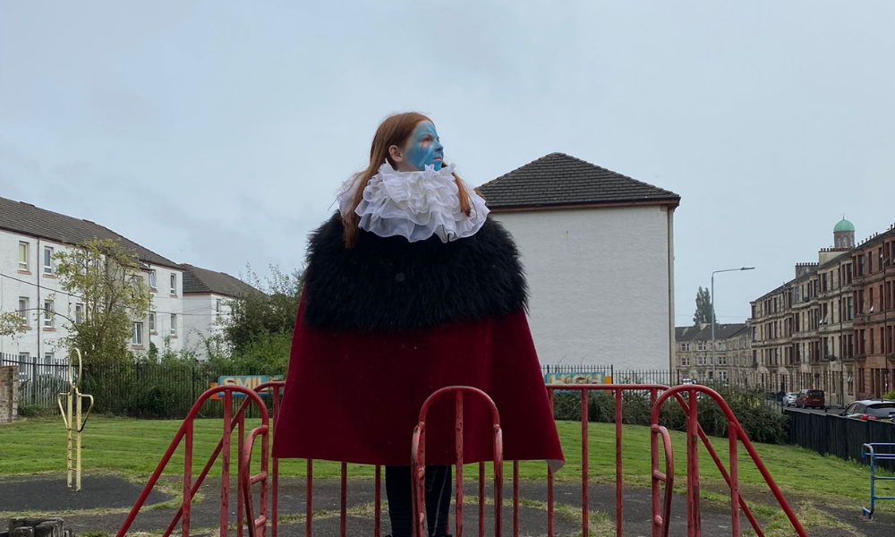 A young participant in a white ruff, black faux fur and red cape and face painted with the Saltire stands at the top of a play park slide.