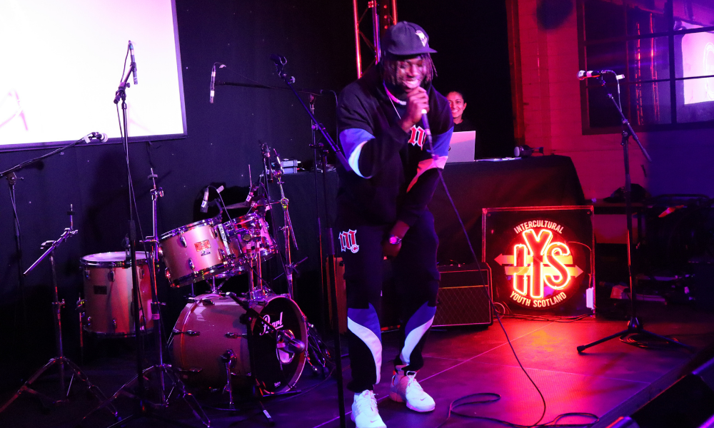CLING performs on a purple lit stage, in front of a drumkit. He holds a microphone and wears a black, blue and white tracksuit and cap with a big smile on his face.