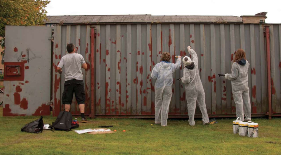 A group of people wearing white overalls paint a metal storage container