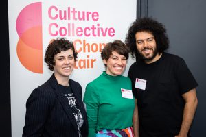 Three people stand in front of a Culture Collective banner, smiling. Morvern, on the left, wears a black smart jacket. Kathryn, in the middle, wears a bright green roll-neck, and Matt, on the right, wears a black t-shirt. 