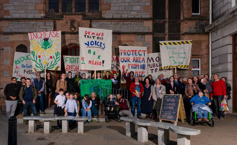 A group of people assemble outdoors holding signs and placards. Two prominent placards read 'Increase biodiversity' and 'Demand that our voices are heard'.